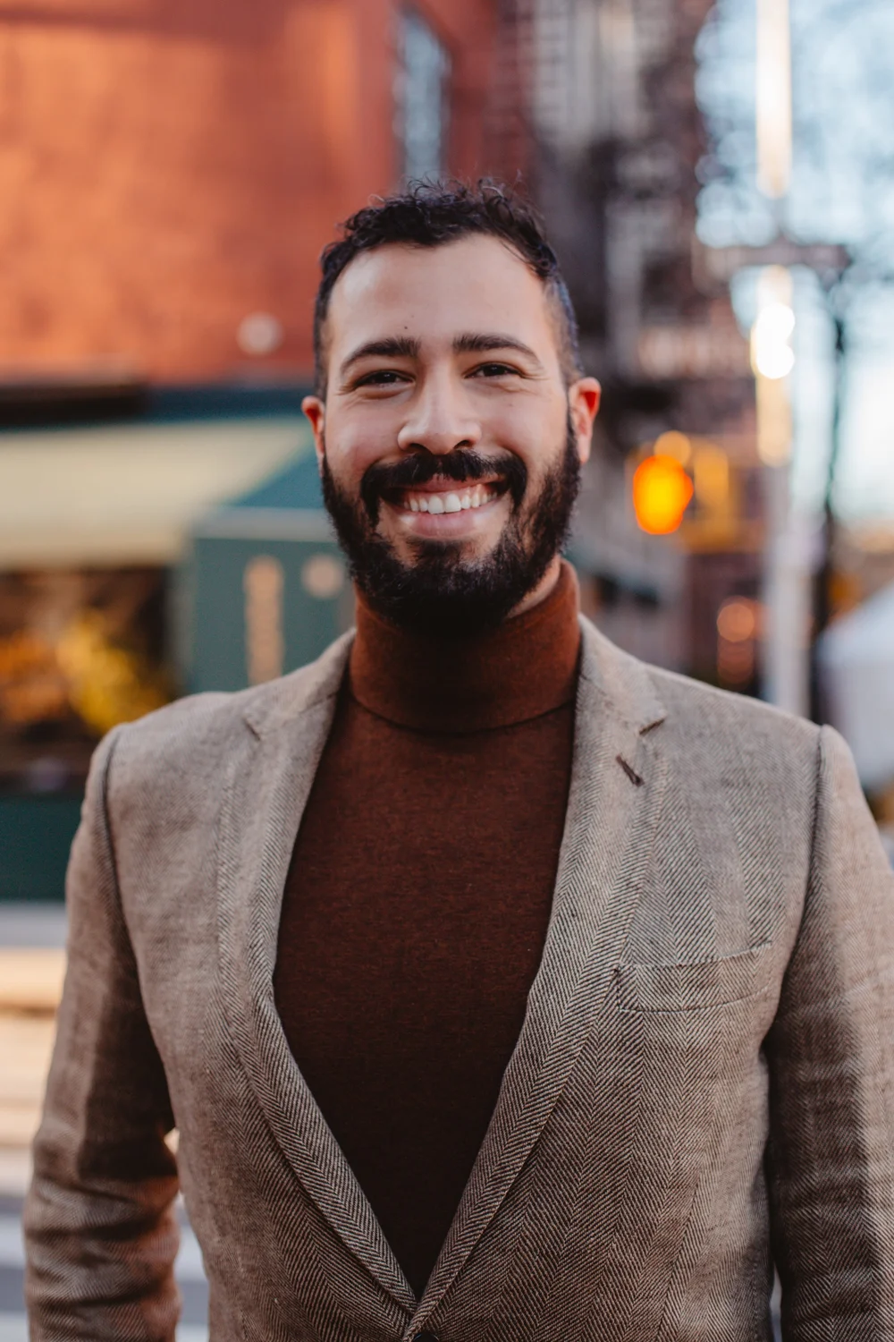 Ramuel Galarza smiling on a city street. He has dark hair and a beard, and wears a tweed blazer over a turtleneck.
